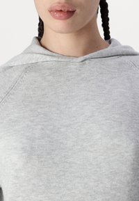 Femme aux cheveux tressés portant un sweat à capuche en tricot gris clair, montrant le décolleté, les lèvres et le haut de la poitrine sur un fond blanc.