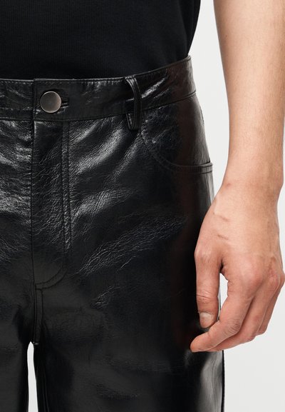 Κοντινό πλάνο ενός ατόμου που φοράει λαμπερό μαύρο δερμάτινο παντελόνι με μεταλλικό κουμπί, δείχνοντας μέρος του χεριού του που στηρίζεται δίπλα στον μηρό.