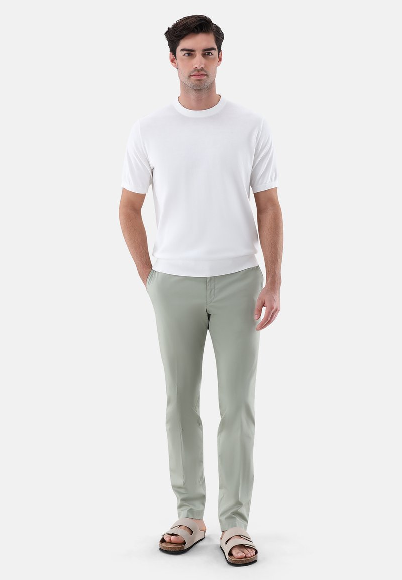 Homme debout portant une chemise blanche à manches courtes, un pantalon gris clair, et des sandales beige à deux lanières, mains détendues, fond neutre.