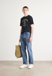 Sort kortærmet t-shirt med gult logo, blå denim jeans, hvide sneakers og en tan totebag, stående på et neutral gulv.