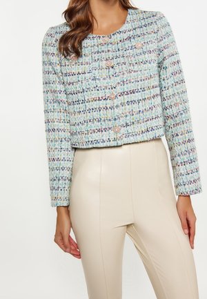 Femme portant une veste courte en tweed multicolore avec des boutons en perle et un pantalon beige taille haute, debout, les mains détendues le long du corps.