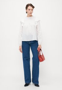 Kvinne iført en hvit bluse med volanger, blå jeans med rett ben, svarte hæler, og holder en liten rød veske med nagledetaljer.