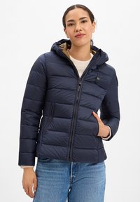 Frau trägt eine marineblaue gesteppte Daunenjacke mit Kapuze, weißen Pullover und blaue Jeans, eine Hand in der Jackentasche.