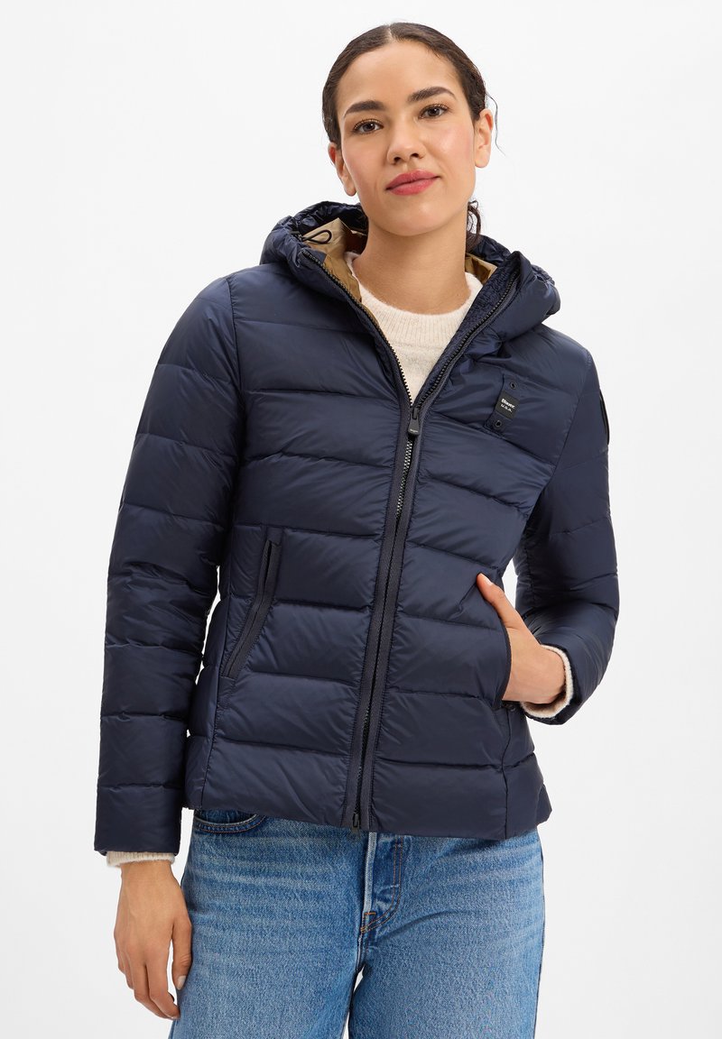 Frau trägt eine marineblaue gesteppte Daunenjacke mit Kapuze, weißen Pullover und blaue Jeans, eine Hand in der Jackentasche.