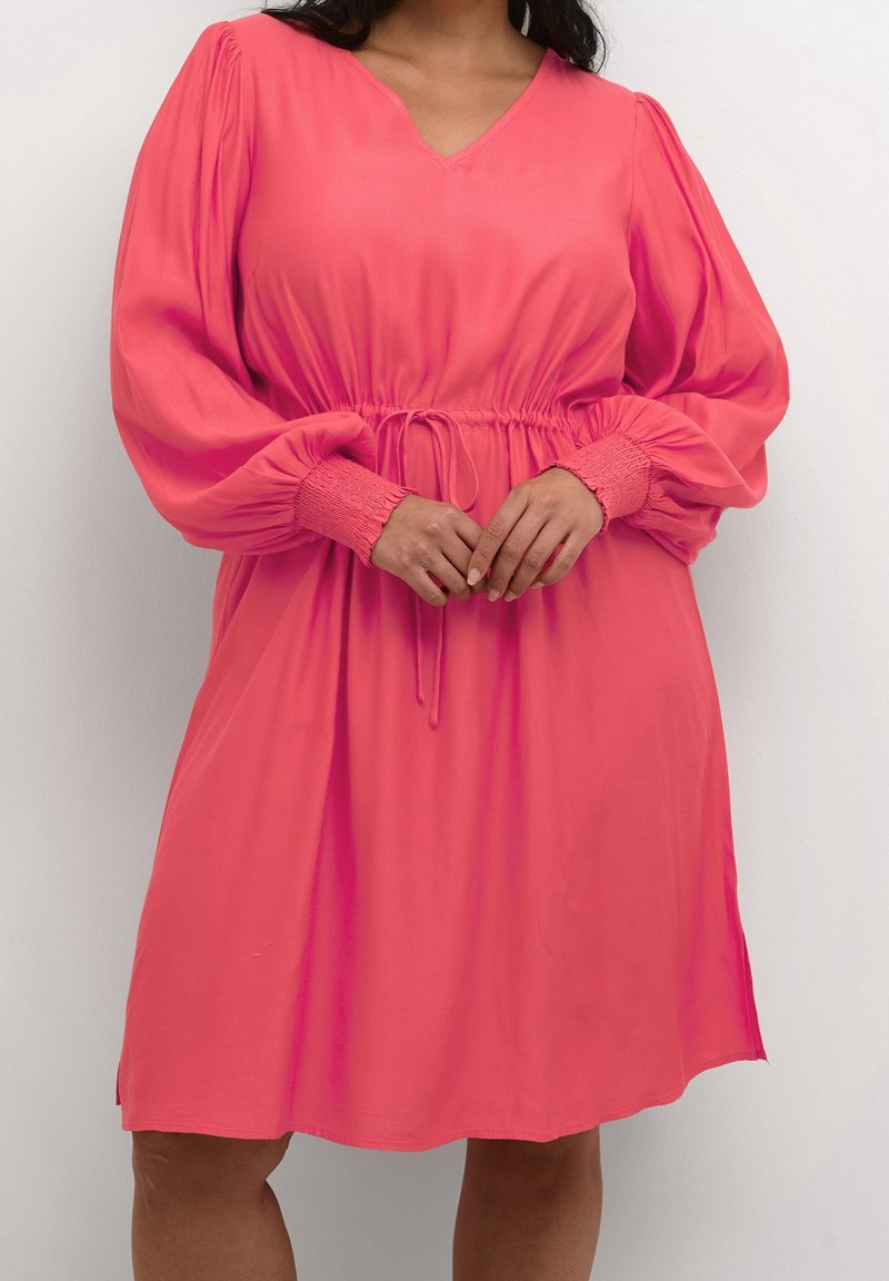Femme portant une robe corail longueur genou avec de longues manches bouffantes, des poignets smockés, une taille cintrée avec un cordon, et un décolleté en V.