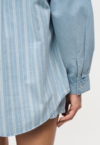 Κοντινό πλάνο ενός ατόμου που φοράει φόρεμα πουκαμίσας με μπλε ρίγες και μανσέτα με κουμπί, δείχνοντας το χέρι και τον μηρό του σε απλό φόντο.