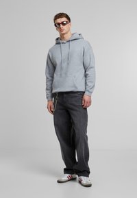 Hoodie vaaleanharmaa, jossa on kengurupussi, yhdistettynä tummiin farkkuihin ja valkoisiin tennareihin, joissa on punaisia yksityiskohtia. Mallilla on aurinkolasit.