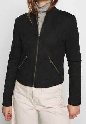 Frau trägt eine taillierte schwarze Jacke mit Reißverschluss und Reißverschlusstaschen und darunter einen grauen, gerippten Rollkragenpullover.