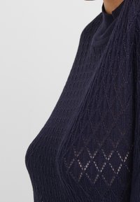 Suéter de punto navy con un patrón de diamantes y un cuello alto, fabricado con un tejido texturizado y transpirable, y una silueta ajustada.