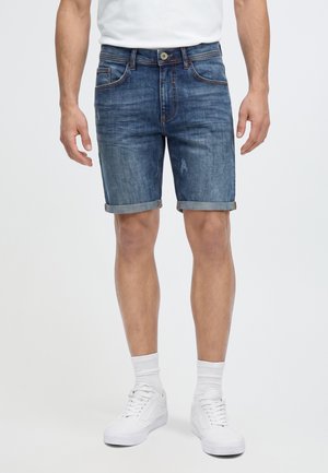 Homme portant un short en jean bleu remonté, des chaussettes blanches, des baskets blanches et un t-shirt blanc sur un fond uni.