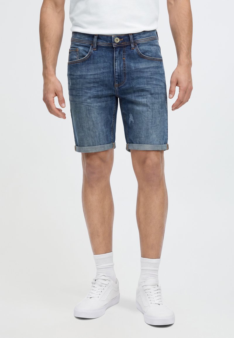 Homme portant un short en jean bleu remonté, des chaussettes blanches, des baskets blanches et un t-shirt blanc sur un fond uni.
