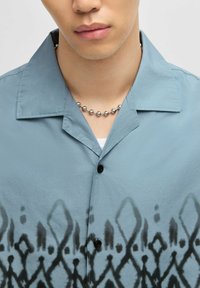 Camicia button-up in tessuto azzurro chiaro, con un motivo geometrico nero lungo l'orlo, dotata di colletto ribattuto e bottoni neri.