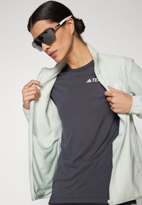 Veste en polaire menthe claire, t-shirt gris avec logo, et lunettes de soleil foncées. La veste présente un zip à l'avant et une texture douce.