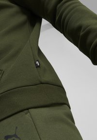 Olivgrön sweatshirt med ribbad mudd, sidoficka och liten svart logotag. Mjuk, texturerad tyg med synliga sömdetaljer.