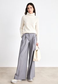 Cremfarbener gerippter Rollkragenpullover, graue plissierte weit geschnittene Hose und eine hellbeige Handtasche. Das Schuhwerk zeigt eine Mischung aus Farben. Minimalistisches Design.