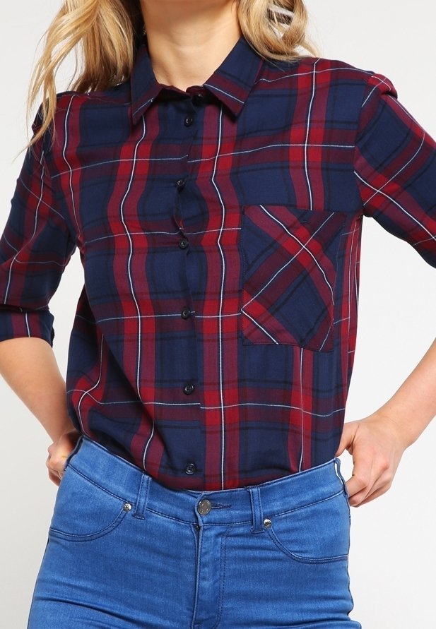 Chemise à carreaux bleu marine et bordeaux avec un devant à boutons, un col et une poche avant présentant des lignes diagonales. Manches longues retroussées.