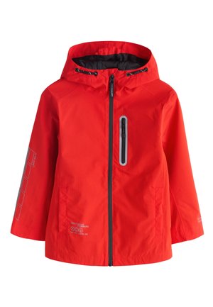 Junge trägt eine leuchtend rote Kapuzenjacke, dunkle Jeans und weiße Sneakers und steht mit entspannten Händen an den Seiten.