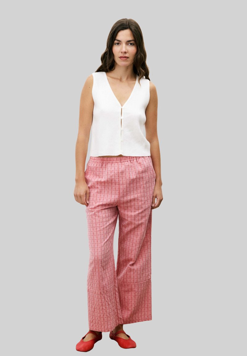 Haut sans manches blanc avec un décolleté en V et des boutons à l'avant, associé à un pantalon large à rayures roses. Des chaussures plates rouges complètent la tenue.