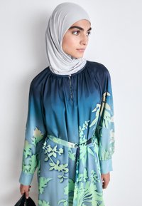 Langes, fließendes Kleid in abgestuften Blau- und Grüntönen mit floralen Mustern. Verfügt über einen gerafften Rundhalsausschnitt und einen gebundenen Gürtel. Ergänzt durch ein hellgraues Hijab.