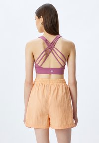 Sostenedor deportivo morado con tirantes entrecruzados; combinado con pantalones cortos naranjas ligeros y elásticos. Textura suave, diseño minimalista y sin logotipos visibles.