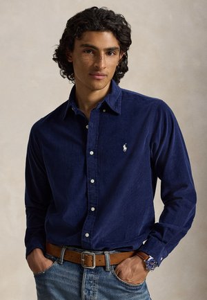 Jeune homme aux cheveux bouclés porte une chemise bleu marine à boutons, une ceinture marron, un jean bleu et une montre-bracelet avec un cadran bleu et un bracelet marron.