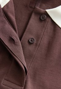 Burgunderfarbene Polo-Shirt mit gerippter Textur, zwei dunklen Knöpfen und einem kontrastierenden weißen Kragen. Nahaufnahme des Kragens und des Knopfleistenbereichs.