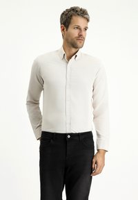 Camicia beige chiaro a maniche lunghe con colletto classico, abbinata a jeans in denim scuri. Tessuto liscio con una leggera trama.