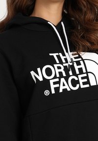 Felpa nera con cappuccio caratterizzata dal logo "THE NORTH FACE" in bianco, con cappuccio regolabile e una texture morbida. Accentuata da un dettaglio circolare del logo.
