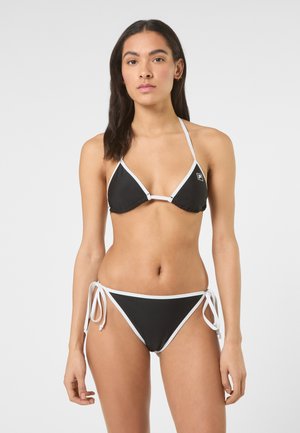 Kvinde iført sort-hvid snøre-bikini med halterneck top og side-bindebukser, stående mod en ensartet lys baggrund.