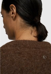 Brauner, flauschiger Pullover mit weicher Textur, der auf der Rückseite ein kleines silbernes Logo zeigt. Der Kragen ist rund und der Stoff wirkt dick.