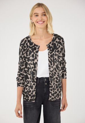 Femme aux cheveux blonds portant un cardigan gris et noir à imprimé léopard sur un top blanc et un jean noir taille haute, souriante.