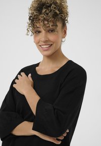 Črna pletena jopica z širokim izrezom in tričetrtinskimi rokavi, ki imajo valovite detajle. Mehka tekstura. Model stoji z zavrčenimi rokami.