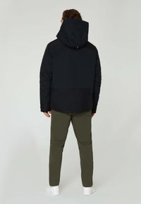 Sort jakke med puffer, der har hætte, og som har et struktureret stof samt et lysere sort panel forneden. Sammen med olivengrønne bukser og hvide sko.