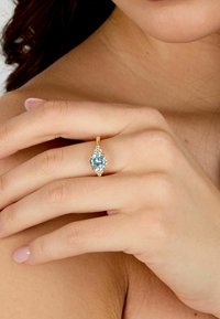 Anello in oro con una pietra blu chiaro a forma di pera, circondata da piccole pietre trasparenti, indossato su una mano con un tono di pelle neutro.