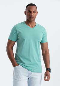 Tričko s výstřihem do V, krátký rukáv, ve světle zelené barvě z bavlny, přiléhavého střihu. Na sobě má světle modré džíny. Modelka stojí s rukama v kapsách.