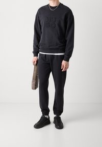 Sudadera negra con el logo "HUGO" en bordado texturizado, pantalones jogger a juego y zapatillas negras. Sosteniendo un pequeño bolso de hombro beige.