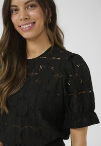 Haut noir avec un design en œillet floral intriqué, manches bouffantes courtes avec des poignets élastiques, tissu texturé et encolure ronde.