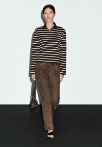 Polo a maniche lunghe a righe in marrone e bianco, abbinato a pantaloni di suede marroni. Il modello porta una piccola borsa marrone scuro e sandali neri.