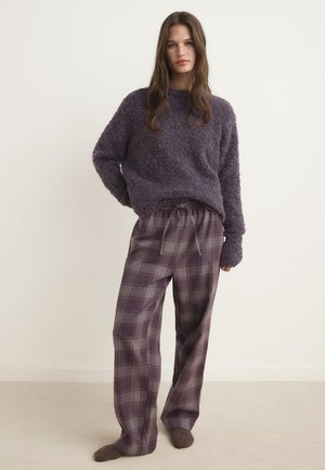 Donna in piedi che indossa un maglione viola peloso, pantaloni a gamba larga a quadri con coulisse e calzini viola coordinati, su uno sfondo chiaro e uniforme.