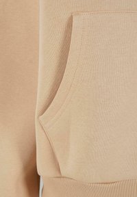 Tessuto di felpa beige con una superficie texturizzata. Presenta una tasca frontale, cuciture visibili e dettagli a coste sull'orlo.