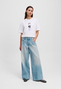T-shirt bianco a maniche corte con un design grafico abbinato a jeans a gamba larga azzurri. Ballerine nere decorate completano l'outfit.