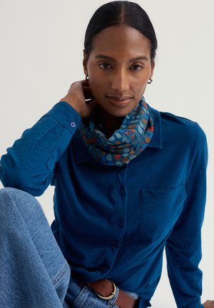 Camicia azzurra con bottoni e taschino, abbinata a una sciarpa decorata con accenti blu, rossi e verdi. Jeans in denim e cintura marrone visibili.