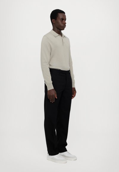 Beige langärmliges Polohemd mit Kragen, kombiniert mit schwarzen Hosen und weißen Sneakern. Das Outfit hat eine lockere Passform und eine glatte Textur.
