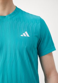 Téglalap alakú textúrájú, diagonális mintázattal rendelkező zöld sportpóló; fehér logó a bal mellrészen; kerek nyakú; rövid ujjas.