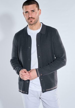Man with short hair and beard wearing dark gray buttoned cardigan over white shirt, posing against light background.