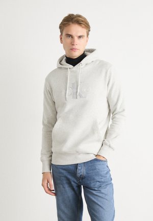 Felpa grigia chiaro con tasca a marsupio, coulisse regolabile e logo ricamato sul petto. Indossata con jeans in denim blu.