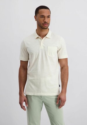 Mann steht und trägt ein off-white kurzärmeliges Polohemd mit Brusttasche und hellgrüne Hose vor einem schlichten Hintergrund.