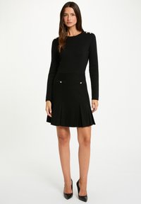 Abito nero con maniche lunghe, corpetto aderente, gonna svasata e pieghe. I bottoni color oro sulle spalle e sulle tasche aggiungono dettagli. Tessuto strutturato.