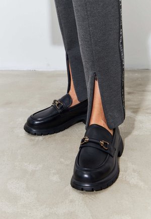 Personne portant un pantalon gris avec des fentes latérales et des mocassins noirs épais avec des détails de boucle dorée, debout sur un sol en béton.