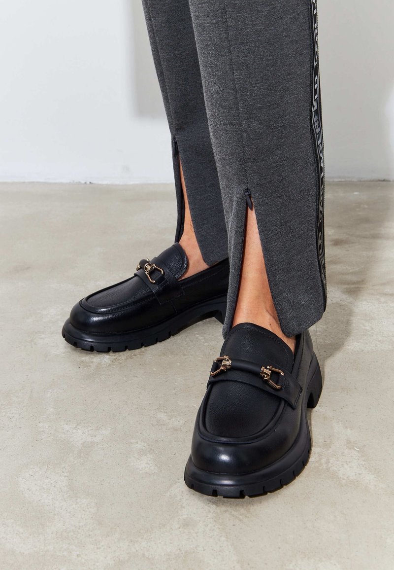 Personne portant un pantalon gris avec des fentes latérales et des mocassins noirs épais avec des détails de boucle dorée, debout sur un sol en béton.